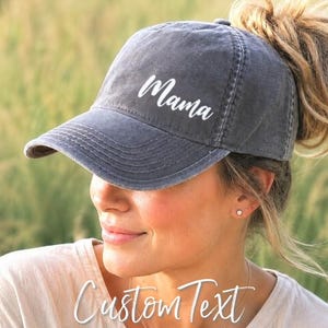 May include: A charcoal gray baseball cap with a distressed, washed-out finish. The word "Mama" is printed in white, cursive script on the front panel. The cap features a curved brim and a structured crown, providing a classic, casual look.