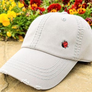 May include: A light gray baseball cap featuring a small embroidered red ladybug on the front panel. The cap has a curved brim with distressed detailing along the edge and matching gray stitching throughout the crown and visor construction.
