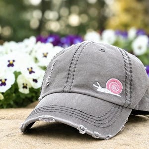 May include: A distressed gray baseball cap with a pink snail embroidery on the front. The cap has a curved brim and decorative stitching. The background features white and purple flowers.