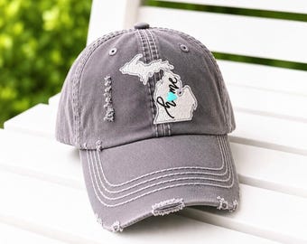 Puede incluir: Una gorra de béisbol gris carbón con aspecto desgastado que presenta un parche blanco bordado del estado de Michigan. La palabra home está cosida en cursiva negra sobre la forma del estado, acentuada por un pequeño corazón turquesa.