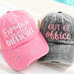 May include: Two distressed trucker hats. One is pink with the text "Expensive and Difficult" in white. The other is gray with the text "OUT OF OFFICE" in pink. Both hats have a mesh back and a curved brim.