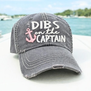 May include: A distressed gray baseball cap with the text "DIBS on the CAPTAIN" in white lettering. A pink anchor graphic is featured on the front. The cap has a pre-worn look with frayed edges.