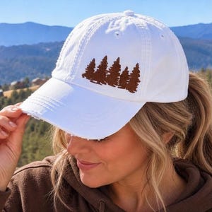 May include: A white baseball cap featuring a brown embroidered graphic of a forest of pine trees on the front panel. The cap has a distressed, frayed brim and a structured crown with ventilation eyelets, providing a classic look.