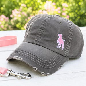 May include: A charcoal gray baseball cap with a distressed, worn-in finish and frayed edges on the brim. The cap features an embroidered patch of a standing dog in light pink with a white outline on the front panel, showcasing a classic six-panel design.