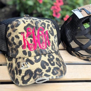 May include: A distressed baseball cap with a leopard print pattern in tan and brown. The cap features a black mesh back and a bright pink embroidered monogram. The brim has distressed detailing.