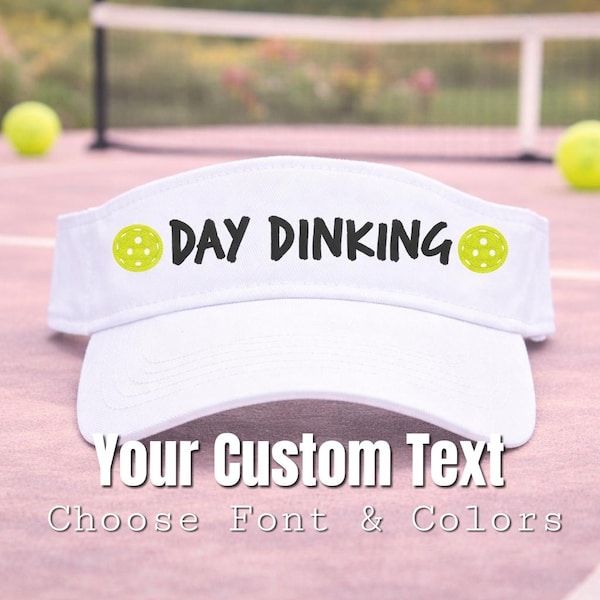 May include: White visor with the text "DAY DINKING" in black, with a pickleball graphic on each side. The visor is on a pink surface with two yellow pickleballs and a net in the background. The text "Your Custom Text" is below the visor.