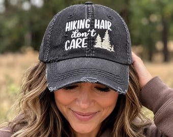 May include: A distressed black baseball cap featuring the white embroidered text HIKING HAIR don't CARE and a small white embroidered graphic of two pine trees on the front panel. The cap has a worn-in look with frayed edges on the brim.