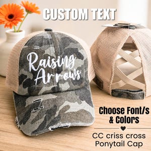 May include: A camouflage-patterned ponytail cap with the text "Raising Arrows" in white script. The cap has a mesh back and a distressed brim. The image also includes text that says "Custom Text" and "Choose Font/s & Colors".