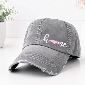 May include: A distressed gray baseball cap with the word "home" in cursive script and a pink outline of North Carolina. The cap has a curved brim and a worn, vintage look.