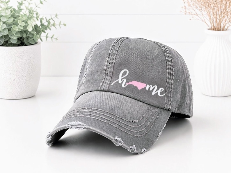 May include: A distressed gray baseball cap with the word "home" in cursive script and a pink outline of North Carolina. The cap has a curved brim and a worn, vintage look.