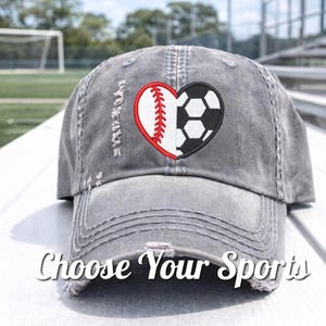 May include: Gray distressed baseball cap with a heart-shaped design featuring a baseball and a soccer ball. The text "Choose Your Sports" is displayed on the front of the cap. The cap is on a white surface.