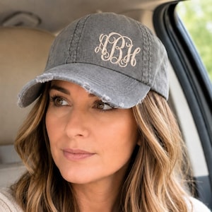 May include: A charcoal gray baseball cap with a distressed, worn-in finish and frayed edges on the brim. The front panel features an embroidered monogram in a light beige, cursive script with the letters ABH intertwined in a decorative style.