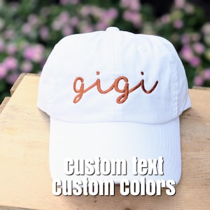 May include: White baseball cap with the word "gigi" embroidered in a copper color. The cap has a curved brim and the text "custom text custom colors" is printed on the front.