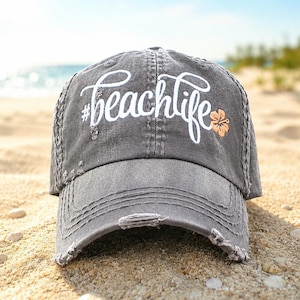 May include: A charcoal gray distressed baseball cap featuring the white embroidered text #beachlife and a small orange hibiscus flower graphic on the front panel. The cap has a worn-in appearance with frayed edges on the brim and seams.