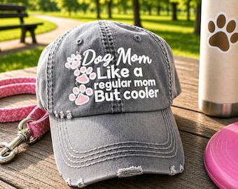 May include: A charcoal gray distressed baseball cap featuring white embroidered text that reads Dog Mom Like a regular mom But cooler, accented with three small pink and white paw print designs on the left side of the front panel.