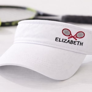 May include: White tennis visor with embroidered crossed rackets and the name "ELIZABETH" in black. Tennis balls and a racket are in the background. The visor is a sporty accessory for sun protection.