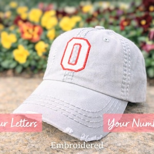 May include: A light gray baseball cap with a distressed look, featuring a red embroidered letter "O". The cap has a curved brim and adjustable strap. The text "Your Letters", "Your Number", and "Embroidered" are also visible.
