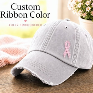 May include: A gray baseball cap with a distressed brim and a pink ribbon embroidered on the front. The text "Custom Ribbon Color" and "FULLY EMBROIDERED" are displayed on the top of the image.