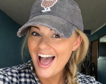 May include: A charcoal gray baseball cap with a distressed, worn-in finish and a frayed brim. The front of the cap features a small, embroidered brown and white chicken graphic with a cross-stitch pattern design.
