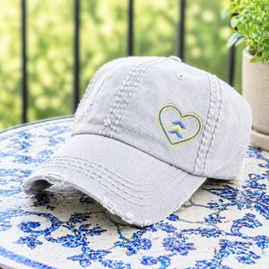 May include: A distressed gray baseball cap with a heart-shaped emblem. The emblem features a yellow outline and two blue arrows inside the heart. The cap has a curved brim and visible stitching details.
