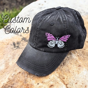 May include: Black distressed baseball cap with an embroidered butterfly in shades of pink, white, and gray. The cap features the text "Custom Colors" in a cursive font. The cap is sitting on a rock.