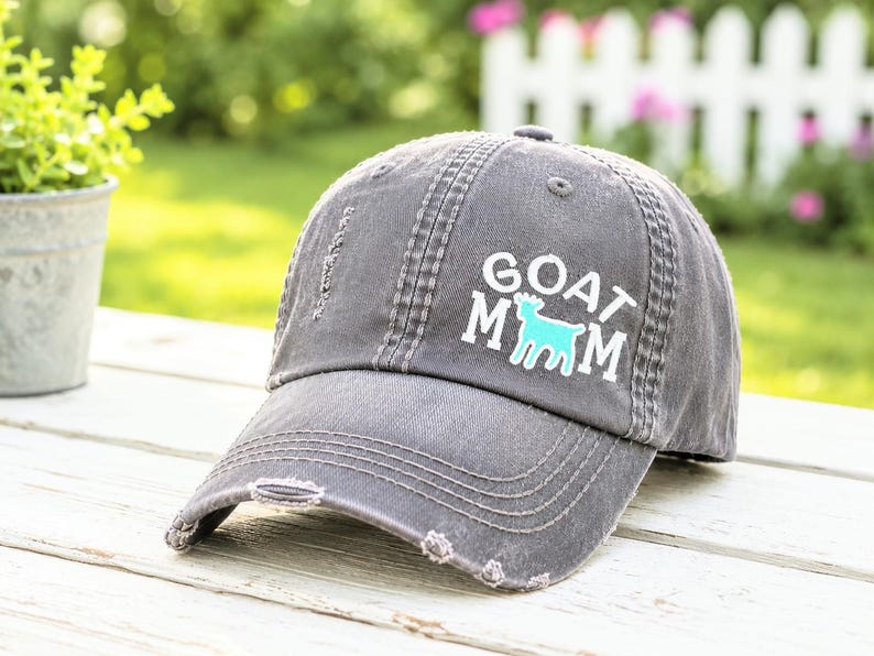 May include: A charcoal gray distressed baseball cap featuring the white text GOAT MOM with a light blue goat silhouette replacing the letter O. The cap has a curved brim with frayed edges and visible stitching details across the front panels.