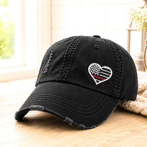 May include: Black distressed baseball cap with a heart-shaped patch. The patch features a design with a white and black checkered pattern, a red stripe, and white stripes. The cap has a curved brim and a worn, vintage look.