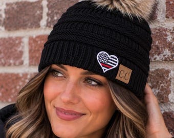 May include: A black knit beanie with a ribbed texture and a large faux fur pom-pom on top. The hat features a heart-shaped patch with a black and white American flag design and a thin red horizontal stripe, plus a small tan C.C. brand tag.