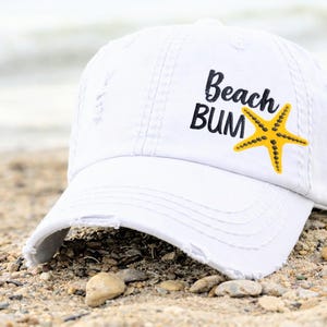 May include: White distressed baseball cap with the text "Beach Bum" and a yellow starfish graphic. The hat is resting on a sandy beach, suggesting a summer or beach theme.