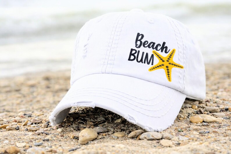 May include: White distressed baseball cap with the text "Beach Bum" and a yellow starfish graphic. The hat is resting on a sandy beach, suggesting a summer or beach theme.