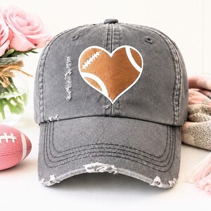 May include: A charcoal gray distressed baseball cap featuring a brown football-shaped heart graphic on the front panel. The cap has a worn, frayed texture along the brim and crown, with a structured six-panel design and a curved visor.