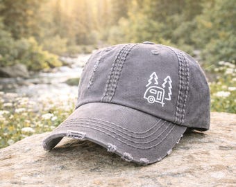 Könnte beinhalten: Graue Baseballkappe im Used-Look mit weißem gesticktem Design eines Wohnwagens und Bäumen. Die Kappe hat einen gebogenen Schirm und sichtbare Nähte. Der Hut liegt auf einem Felsen, mit verschwommenem Naturhintergrund.