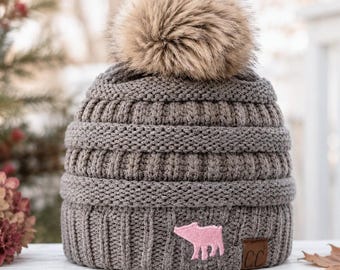 May include: A grey knit beanie featuring a thick ribbed texture and a large, fluffy tan faux fur pom-pom on top. The hat includes a small pink embroidered pig patch on the front cuff and a brown rectangular tag with the letters CC embossed.