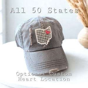 May include: Gray distressed baseball cap with a patch featuring the state of Ohio with a red heart. The cap has the text "All 50 States" and "Optional Custom Heart Location".