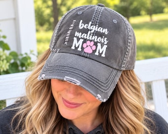 May include: A charcoal grey distressed baseball cap featuring the white text "belgian malinois" above the word "MOM" in white block letters, with a pink paw print icon replacing the letter "O" in the centre of the design.
