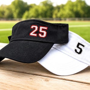 May include: Two visors, one black and one white, rest on a wooden surface. The black visor displays the number "25" in white with a red outline. The white visor has the number "5" in black with a white outline. The background is a green field.