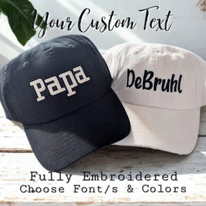 May include: A dark charcoal baseball cap with the white embroidered text Papa and a cream baseball cap with the black embroidered text DeBruhl. The image includes the text Your Custom Text and Fully Embroidered Choose Font/s & Colors.