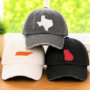 May include: Three distressed baseball caps in gray, white, and black. Each cap features a state outline: Texas in white on gray, Tennessee in orange on white, and Georgia in red on black. The caps have a worn, vintage look.