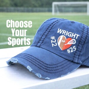 May include: A navy blue distressed baseball cap featuring white embroidered text that reads WRIGHT, #22, and #25, alongside a heart-shaped graphic containing a football and a lacrosse stick. The text Choose Your Sports is visible on the left.