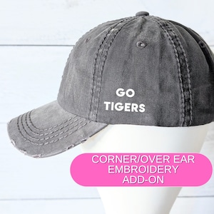 May include: A gray distressed baseball cap with white embroidery that reads "GO TIGERS". The cap is shown on a white mannequin head. The text "CORNER/OVER EAR EMBROIDERY ADD-ON" is displayed in pink on a white background.