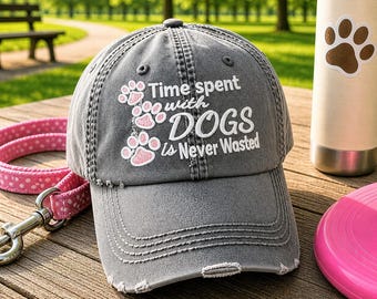 May include: A charcoal gray distressed baseball cap featuring white embroidered text that reads Time spent with DOGS is Never Wasted, accented by three small pink paw prints arranged vertically on the left side of the front panel.