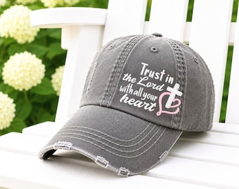 May include: A charcoal gray distressed baseball cap featuring the white text "Trust in the Lord with all your heart" alongside a white cross and a light pink heart outline printed on the front panel of the hat.