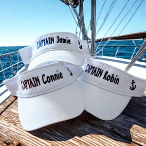 May include: Three white visors with the text "CAPTAIN Jamie", "CAPTAIN Connie", and "CAPTAIN Robin" embroidered in blue and maroon thread. Each visor has a small black anchor symbol. The visors are on a wooden surface with a blue ocean background.