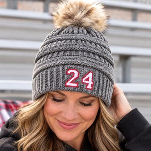 May include: A charcoal gray cable-knit beanie featuring a thick ribbed cuff and a large, fluffy faux fur pom-pom on top. The front of the cuff displays the number 24 in white digits with a red outline, stitched onto a rectangular patch.