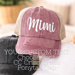 May include: A collection of distressed baseball caps in various colors, including a dusty rose cap with "Mimi" in white script. The caps have mesh backs and are designed for a ponytail. Text on the image reads "YOUR CUSTOM TEXT Choose Font CC Criss Cross Ponytail Cap."
