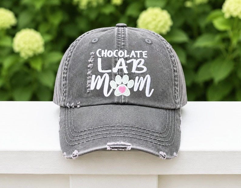 Pu&ograve; includere: Un berretto da baseball grigio antracite dal look vissuto, con la scritta bianca "CHOCOLATE LAB MOM" e una grafica a forma di zampa bianca con un piccolo cuore rosa al centro. Il berretto presenta una finitura usurata con bordi sfilacciati.