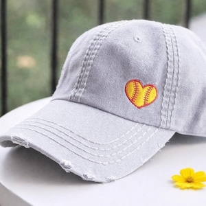 May include: A light gray baseball cap with a distressed brim and stitching details. The cap features an embroidered yellow and red heart-shaped softball design on the front. A small yellow flower rests on the surface.