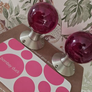 May include: Two spherical, deep red glass door knobs with brushed nickel bases. They rest on a brown surface beside a card with pink circles and the text "bombay duck". Floral wallpaper forms the backdrop.