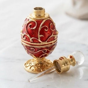 Op de afbeelding: Een eivormig parfumflesje met een levendige rode emaille afwerking en ingewikkeld goudkleurig filigraan krulwerk. Het flesje rust op een ronde goudkleurige voet en bevat een bijpassende goudkleurige dop met een glazen applicatorstaafje.