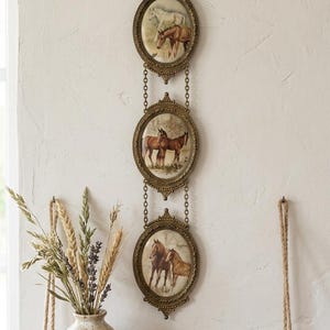 Puede incluir: Un colgante de pared vertical con tres marcos ovalados de latón conectados por cadenas, cada uno con una ilustración de caballos. Los marcos cuelgan de un solo gancho de latón. Las obras de arte representan varios caballos en exteriores.